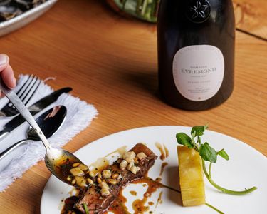 A bottle of Domaine Evremond English quality sparkling wine is paired with a gourmet plated dish featuring finely presented meat, sauce, and vegetable accompaniments on a wooden table. Elegant dining cutlery and a flower vase add to the refined setting, emphasizing premium craftsmanship and the vineyard-inspired culinary experience associated with Domaine Evremond.